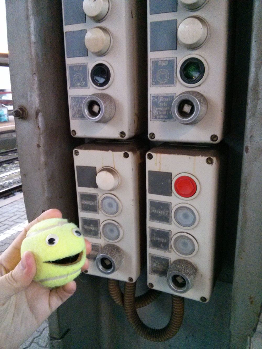 Ball-kun hält einen grünen Tennisball vor alten Telefonzellen.