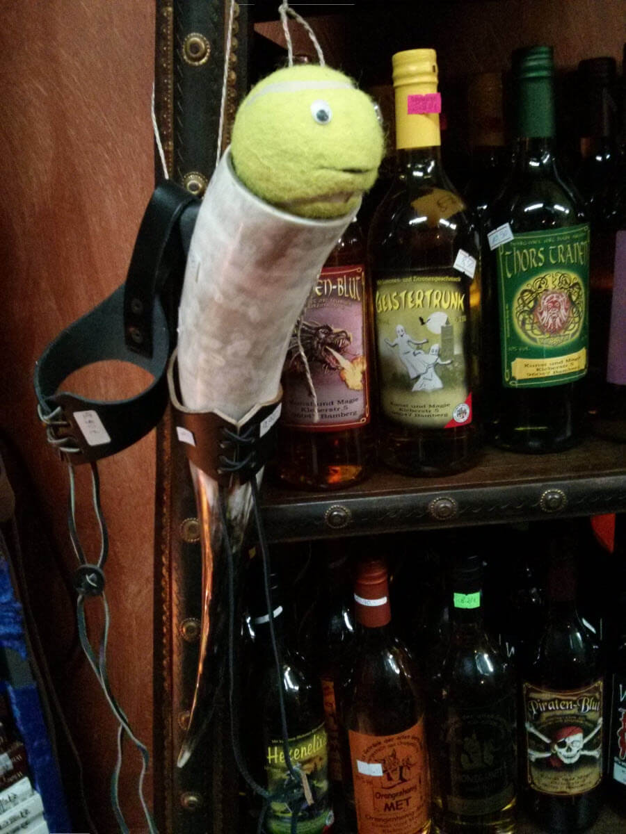 Plüsch-Fisch mit Ball-Kun Kopf hängt an Wand, Weinflaschen im Hintergrund.