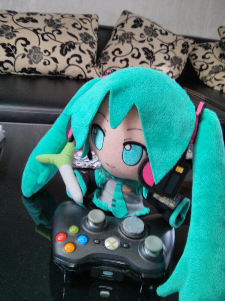 Hatsune Miku Plüschfigur mit Controller auf Tisch, Couch im Hintergrund.