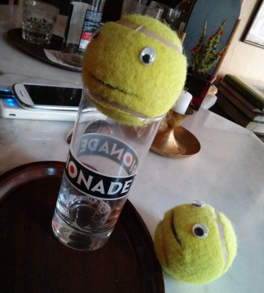 Tennisball mit Augen auf einem Glas, daneben ein weiterer Tennisball.
