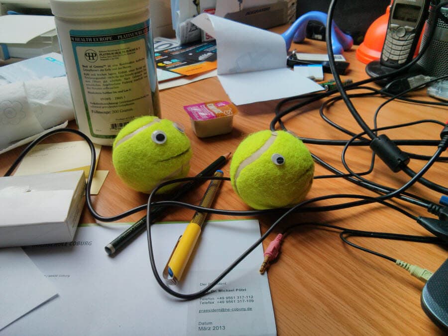 Zwei grüne Tennisbälle mit googly eyes auf einem chaotischen Schreibtisch.