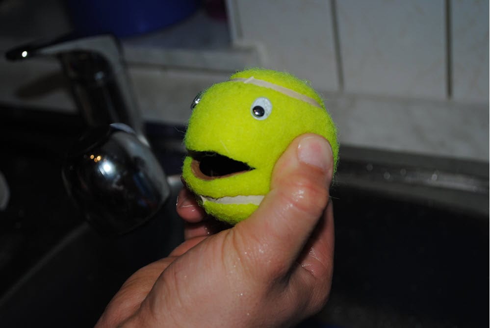 Gelber Ball mit Augen und Mund, gehalten in Hand über Wasserhahn.