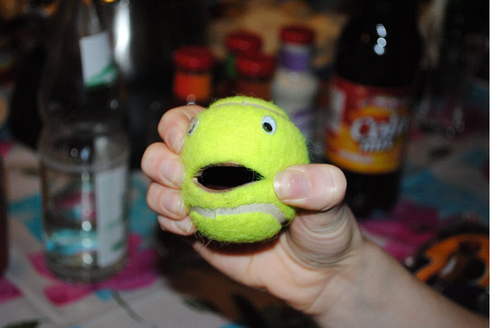 Hand hält einen grünen Ball mit Gesicht, Hintergrund mit verschiedenen Gegenständen.